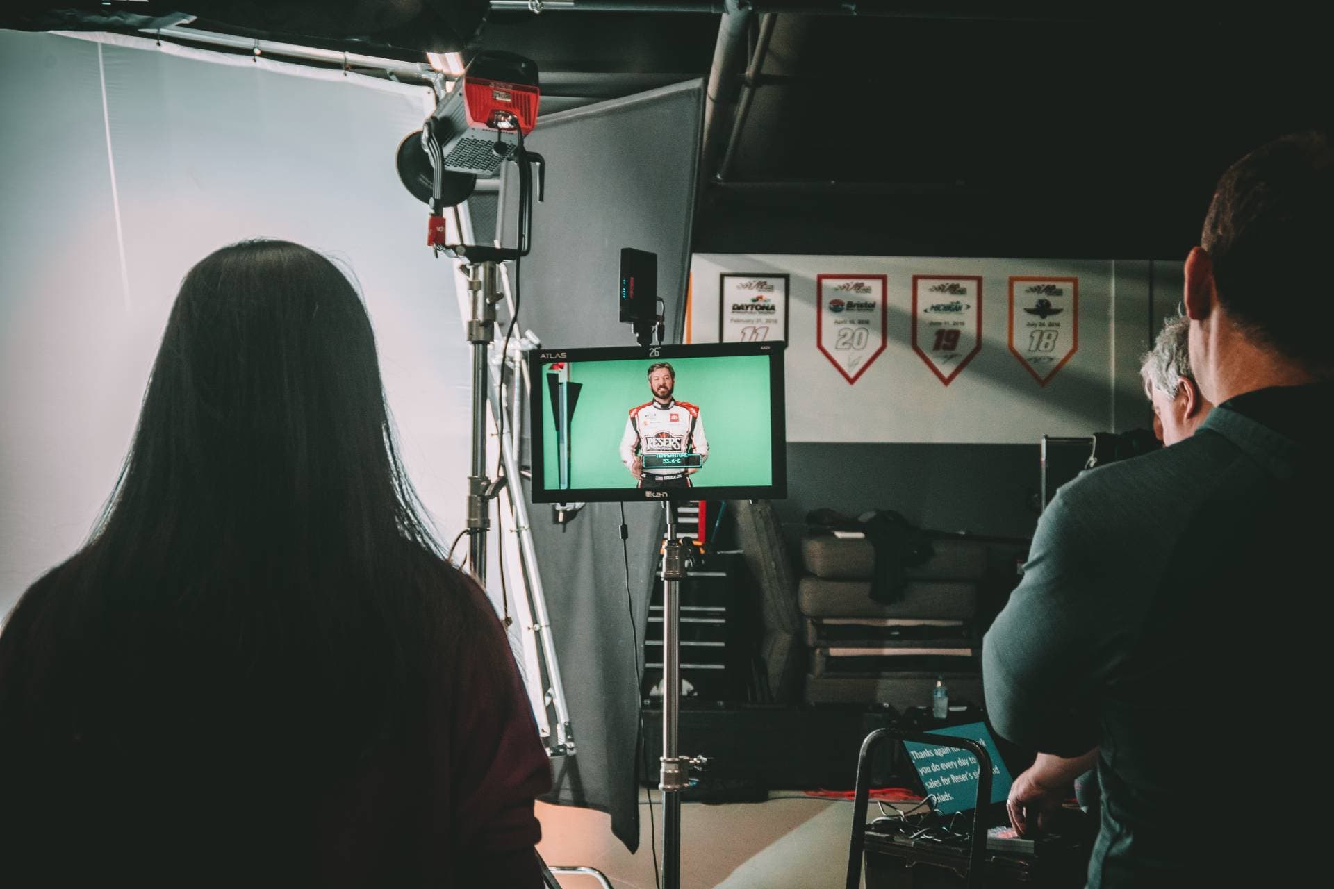 Crew watching a monitor during a video shoot — a driver in a branded racing suit filmed against a green screen in a race team's shop